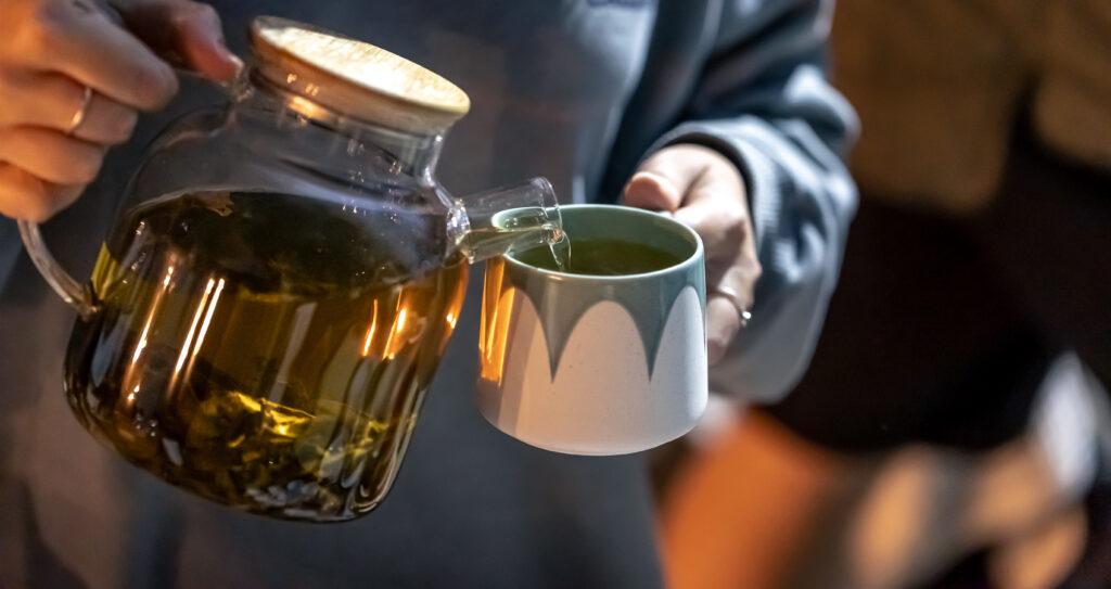 A woman pours tea infusion into a cup with herbs.